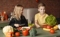 Positive little girl and boy sitting at table with assorted vegetables and fruits while preparing healthy food together in loft kitchen with brick wall